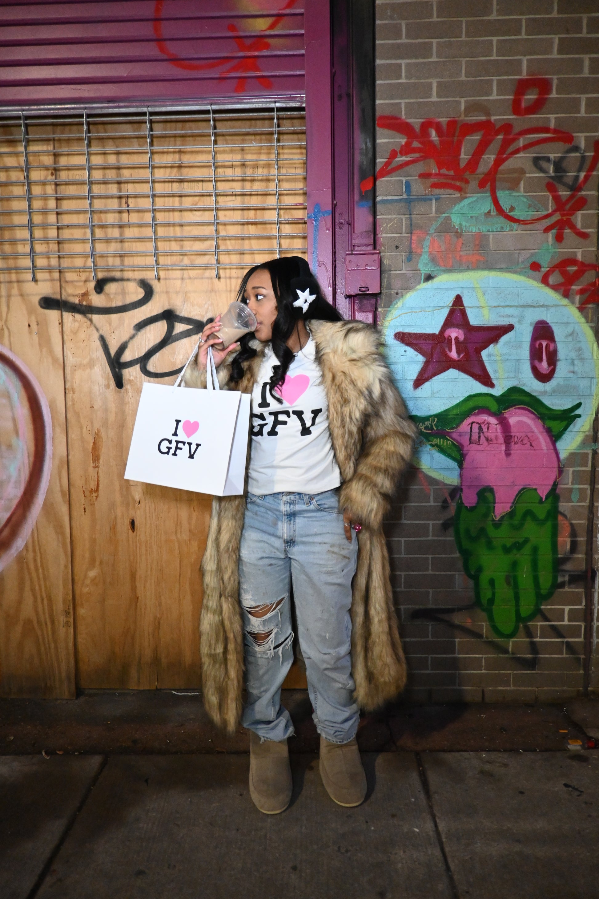 Person standing on a street drinking coffee in an I heart gfv t shirt holding a shopping bag, surrounded by graffiti on the sidewalk.